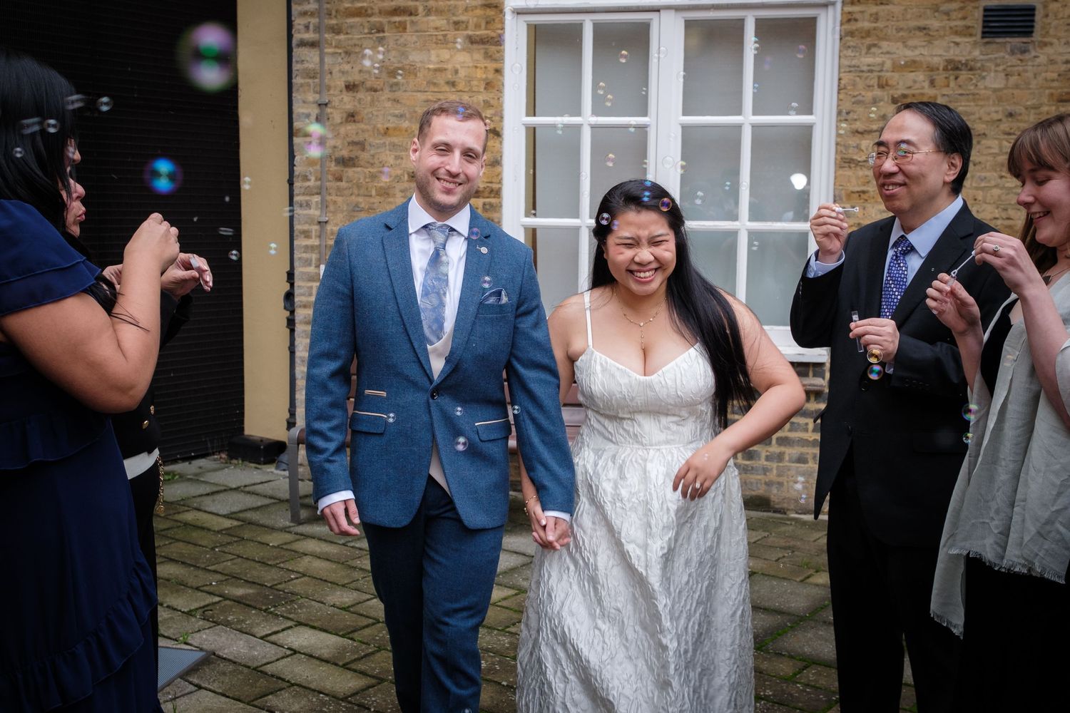 Bride and groom walk hand in hand as guests blow bubbles during outdoor wedding celebration at brick venue.