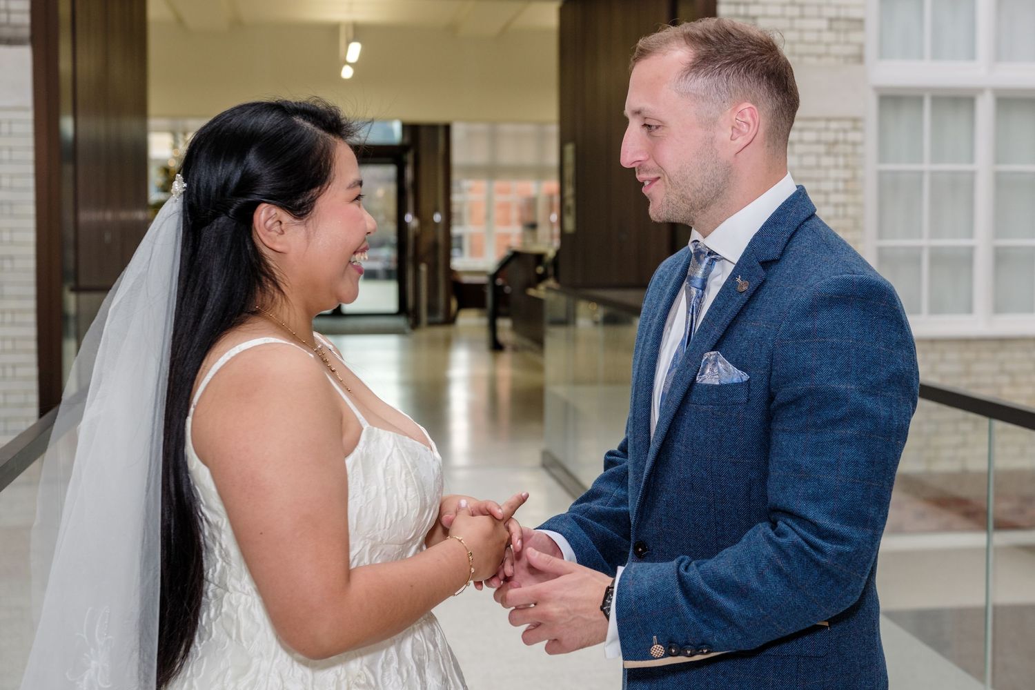 Bride and groom holding hands and smiling at each other during wedding ceremony in elegant indoor venue.