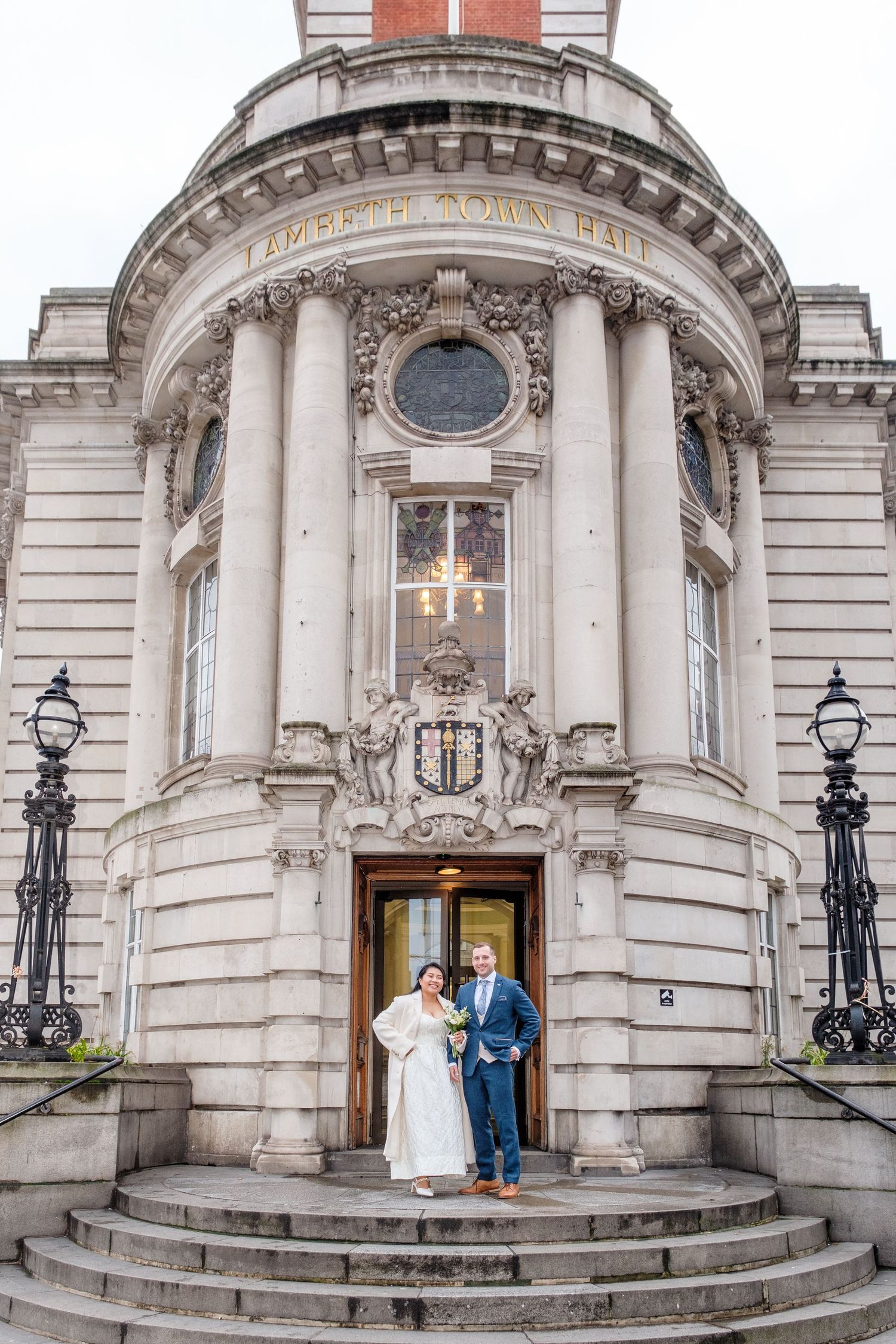 Bride and groom posing on steps of Lambeth Town Hall, a grand neoclassical building with columns, in London.