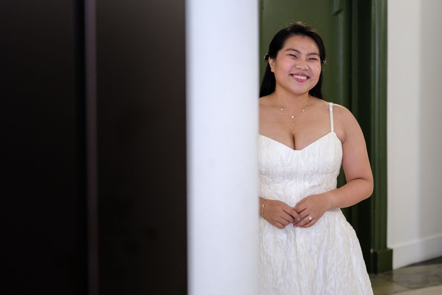 Smiling bride in a white lace wedding dress standing by a white pillar in an elegant indoor setting.