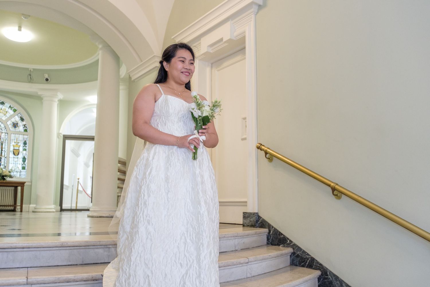 Bride in white wedding gown holding bouquet stands on steps inside elegant venue with arched windows.