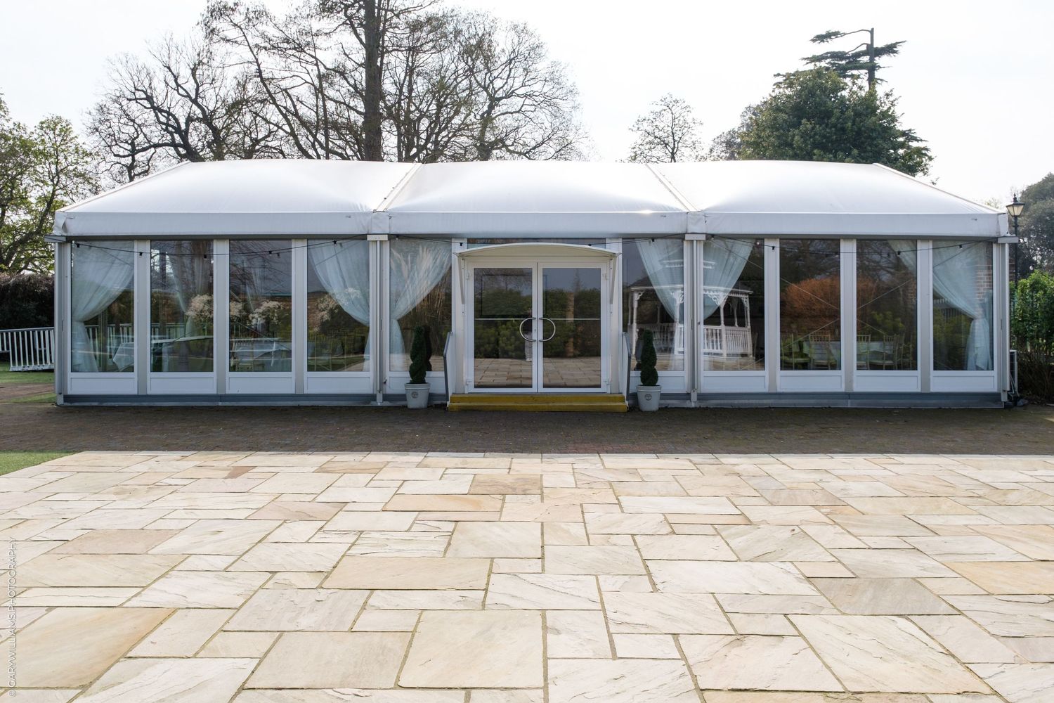Large white marquee tent with glass walls and white curtains set on a stone patio surrounded by trees.