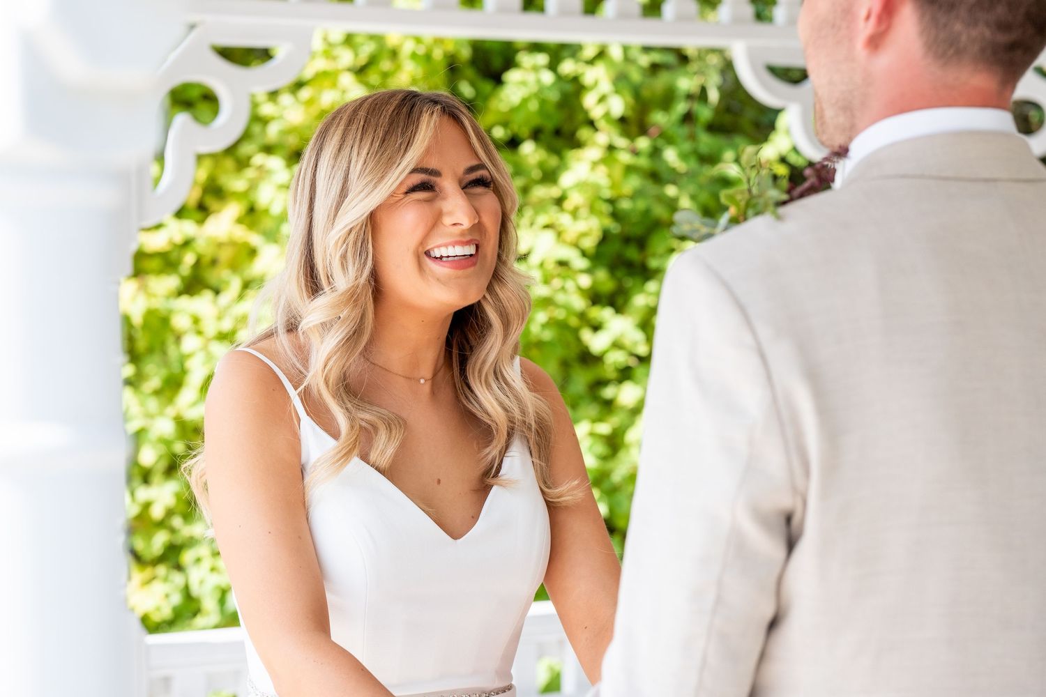 Smiling bride in white dress stands at outdoor wedding ceremony under white pergola with lush green trees.