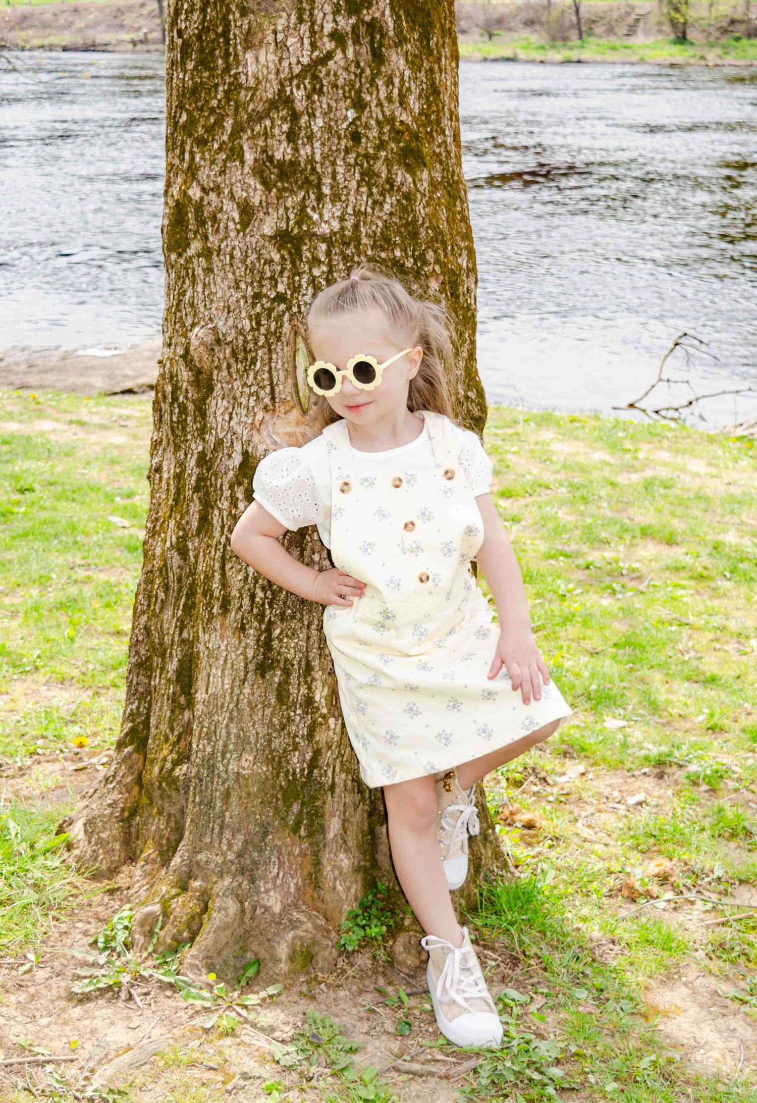 Baby in white dress with glasses sits in wooden crate beside FRAGILE box and lamp, Christmas tree background.