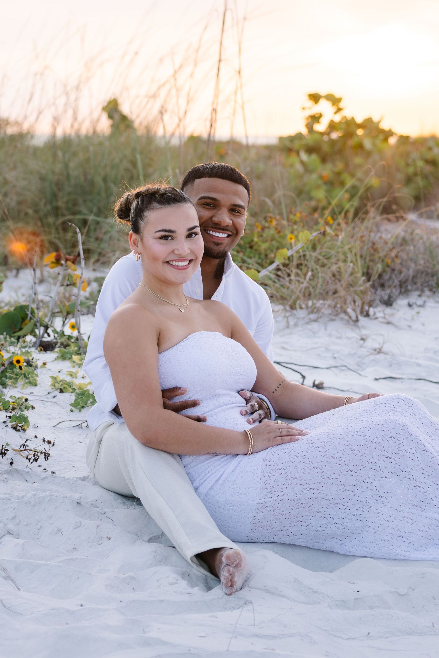 A smiling couple in white outfits sits on sandy beach at sunset, the woman leaning back against the man.