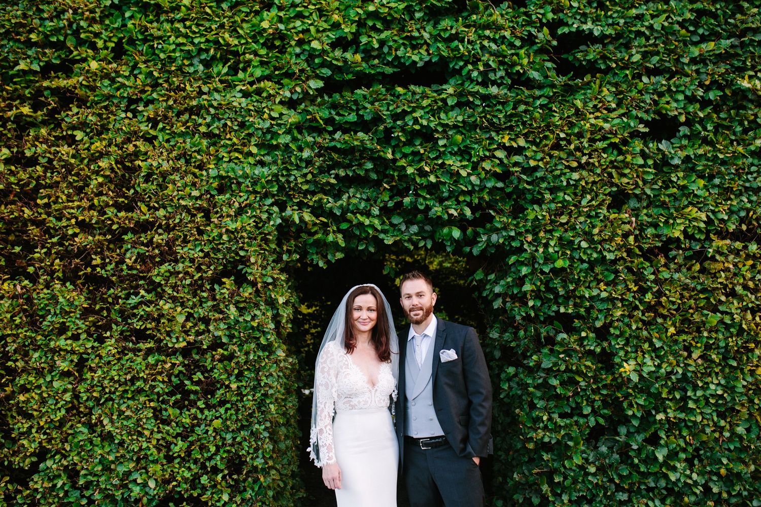 Bride and groom standing together in front of a lush green ivy hedge wall at an outdoor wedding venue.