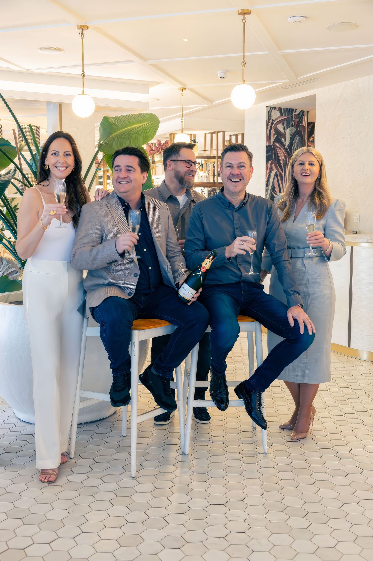 Five professionals celebrating with champagne glasses in a modern, bright interior space with hexagonal tile floors.