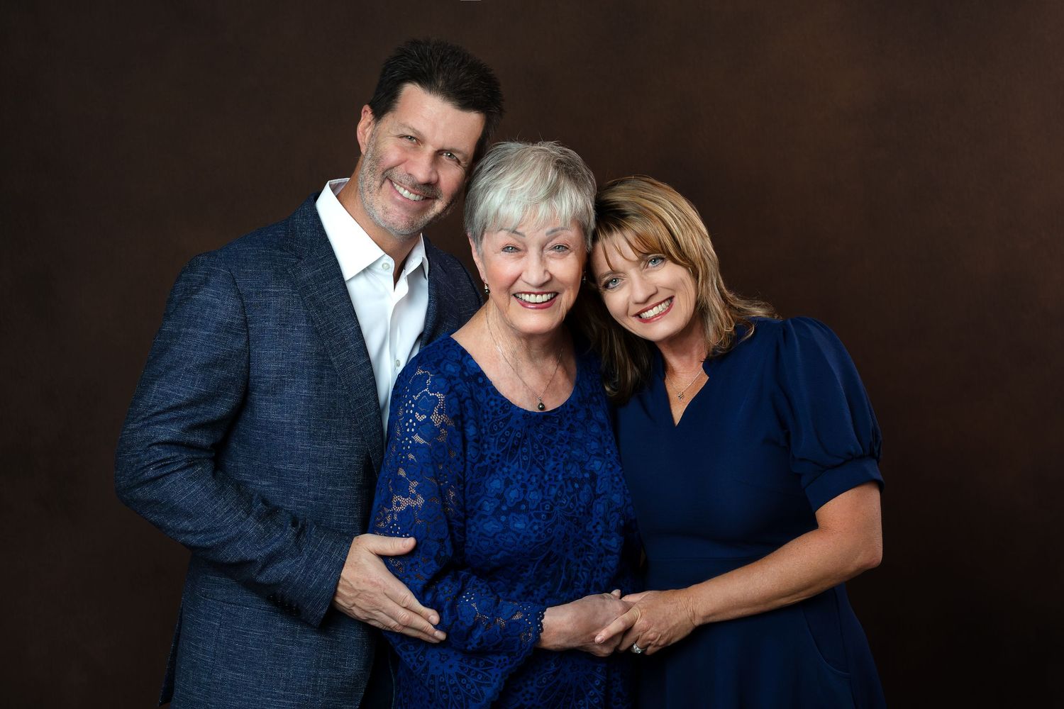 Three smiling adults pose together in coordinating blue outfits against a warm brown studio backdrop.