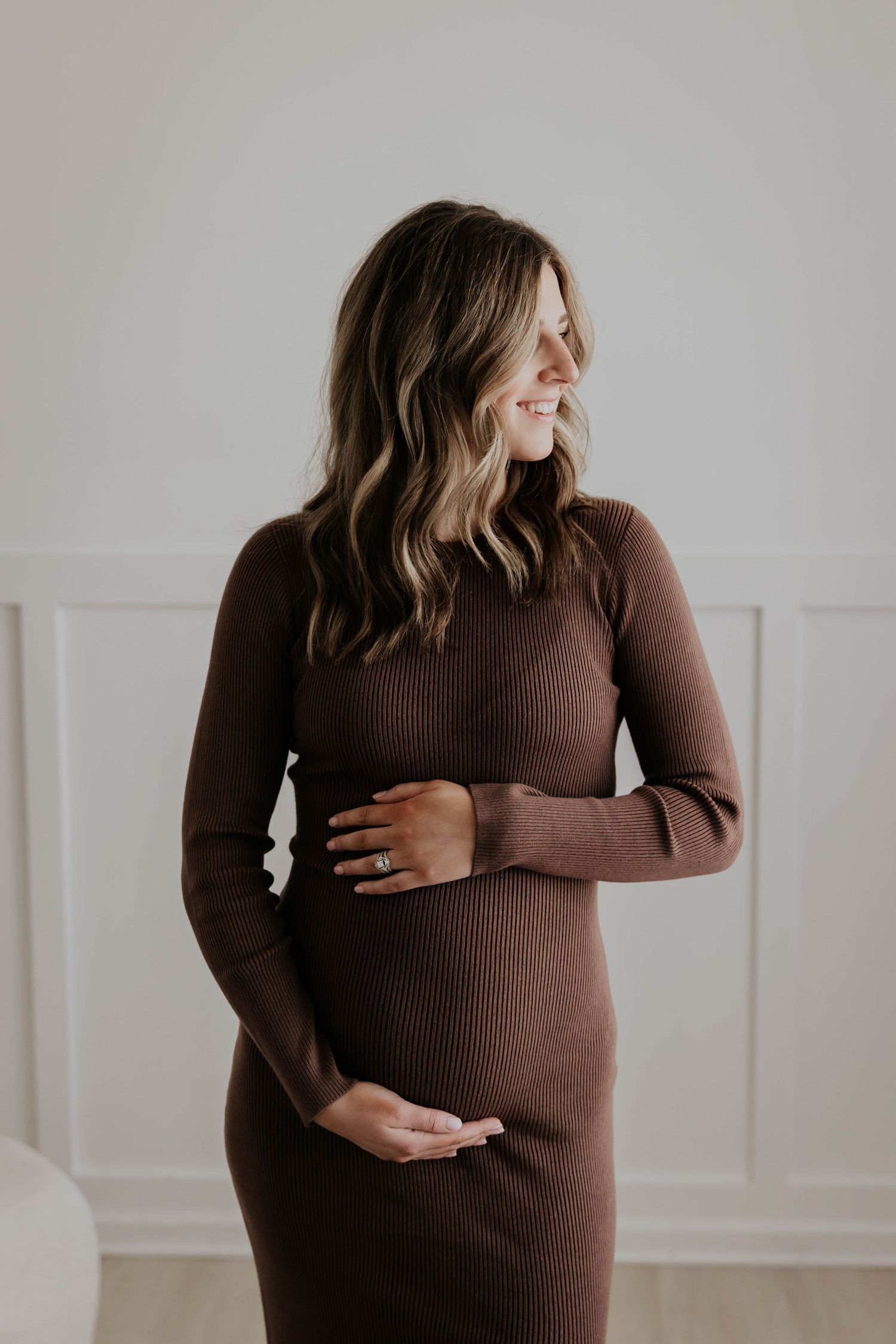 Pregnant woman in white shirt and jeans cradling baby bump near large windows with brick wall background.