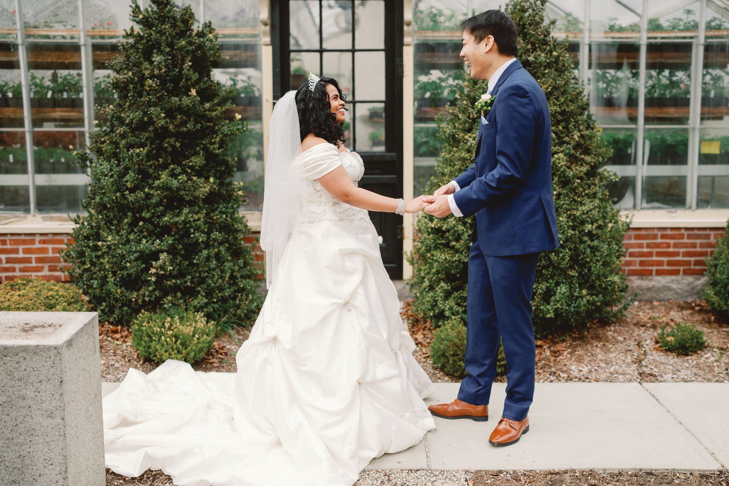 Bride in white gown and groom in navy suit hold hands outdoors near evergreen trees at elegant wedding venue.