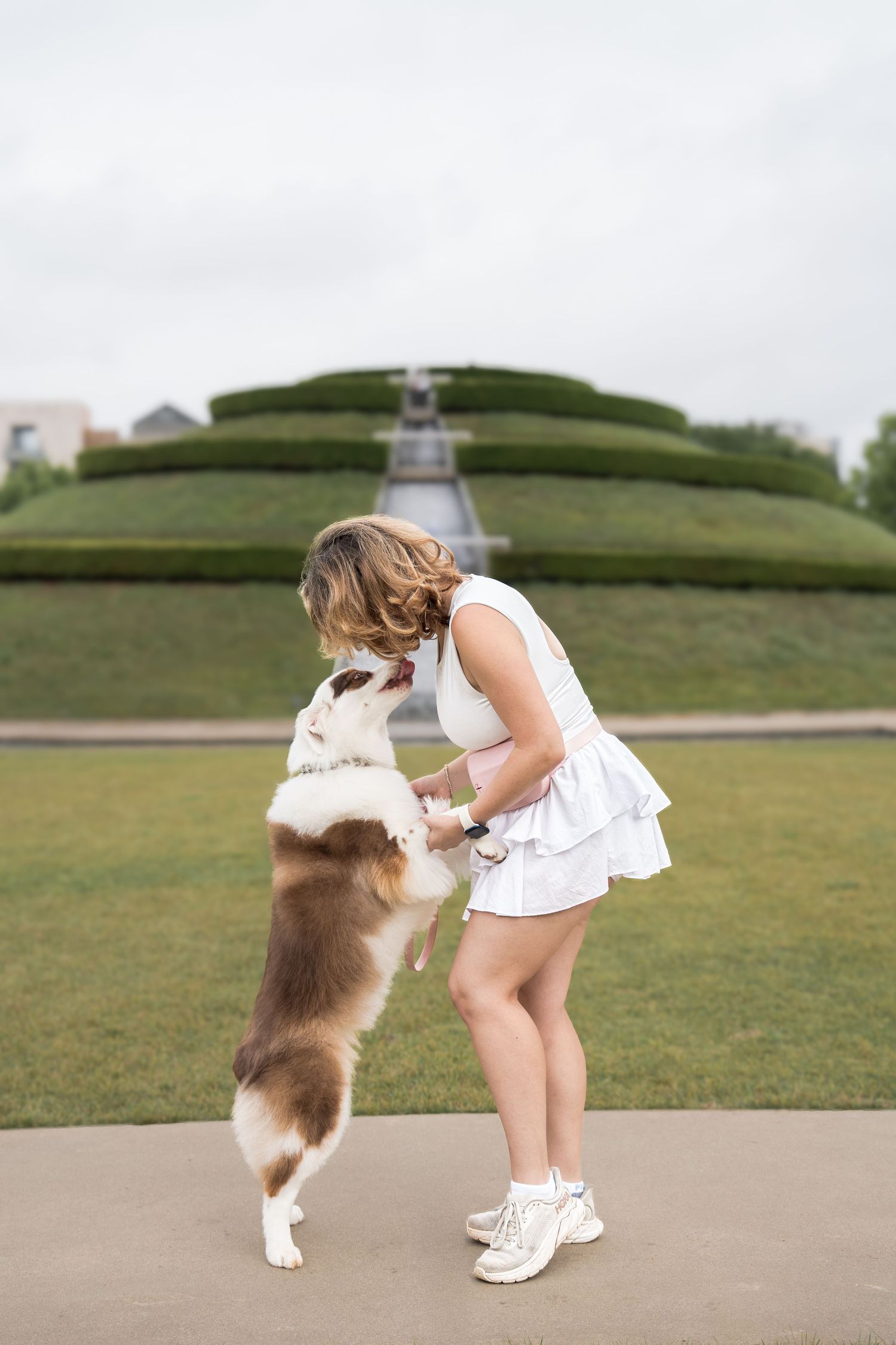 Woman in white dress playing with jumping Australian Shepherd dog on green lawn outdoors.