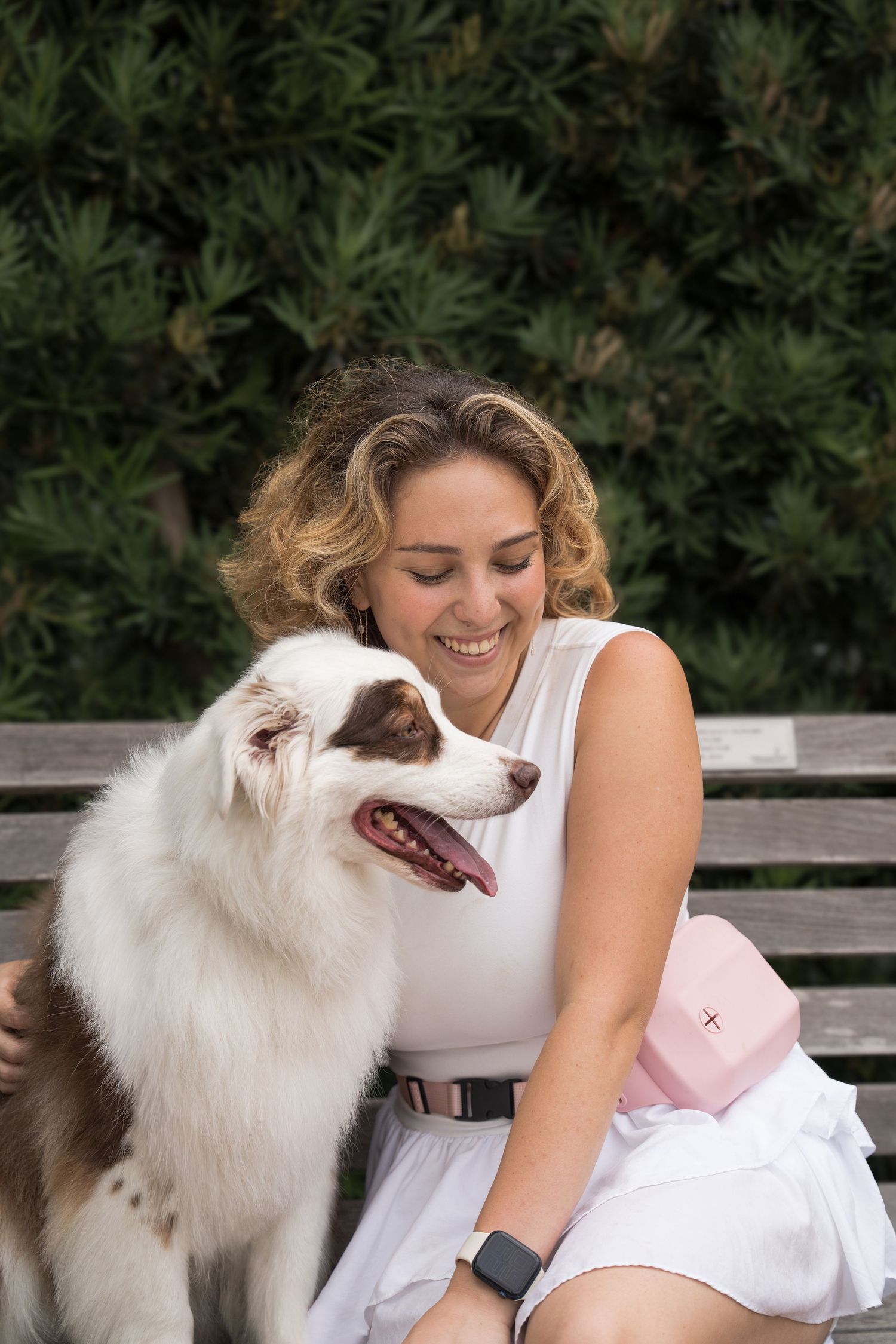 Happy woman in white outfit hugs fluffy brown and white dog on park bench.