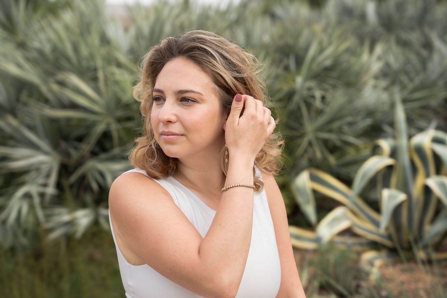 Woman with curly blonde hair in white top adjusting her hair outdoors near tropical plants.