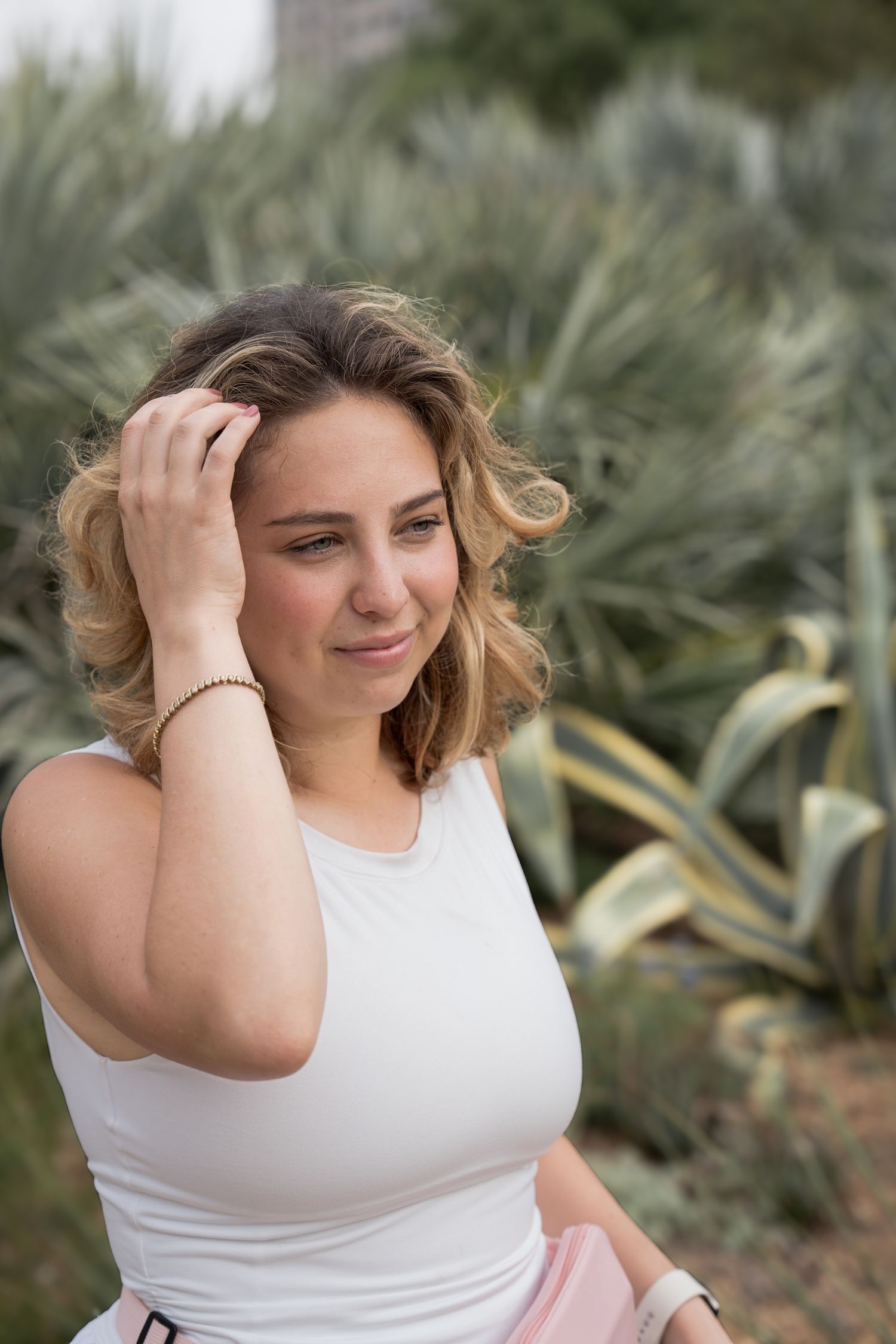 Young woman with curly blonde hair in white top, posing outdoors near green agave plants.