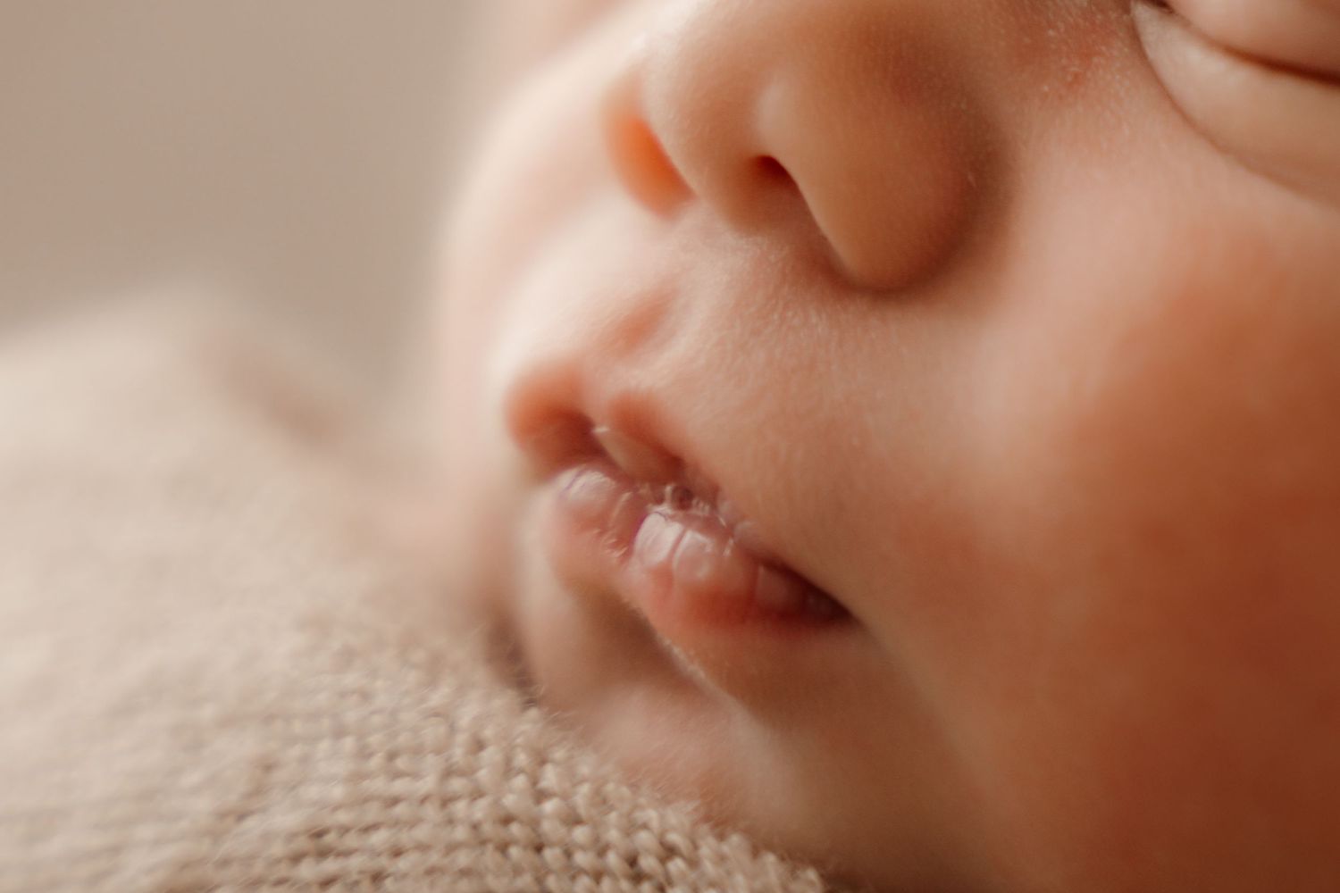 Close-up macro photo of a sleeping newborn baby's nose and lips on a soft beige knit blanket.
