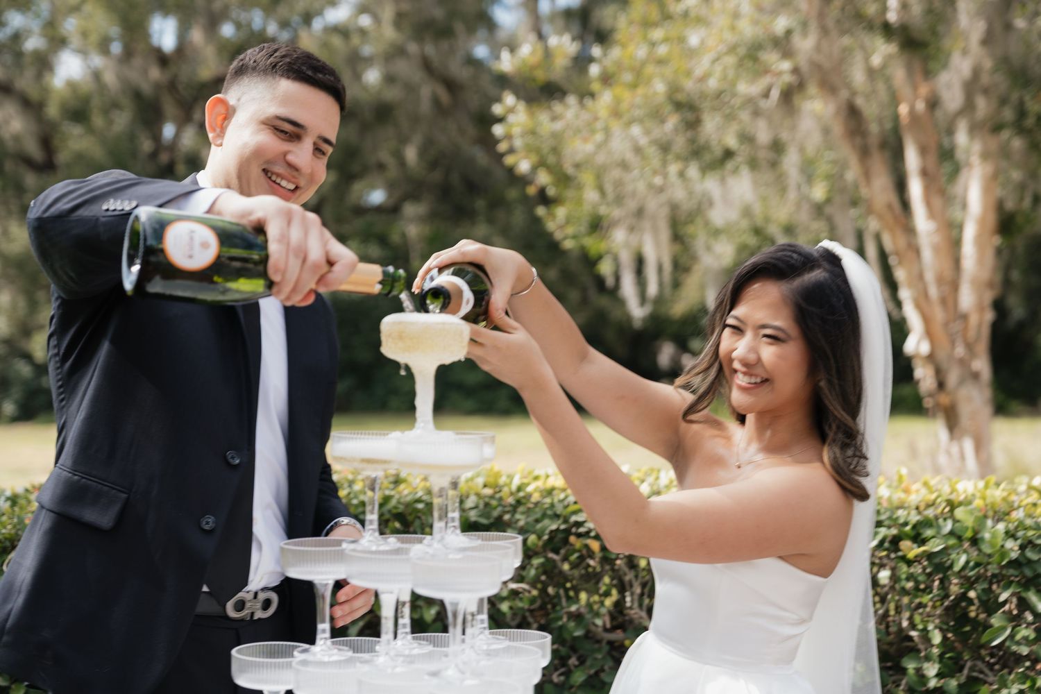Bride and groom pouring champagne tower at outdoor wedding celebration surrounded by lush greenery.
