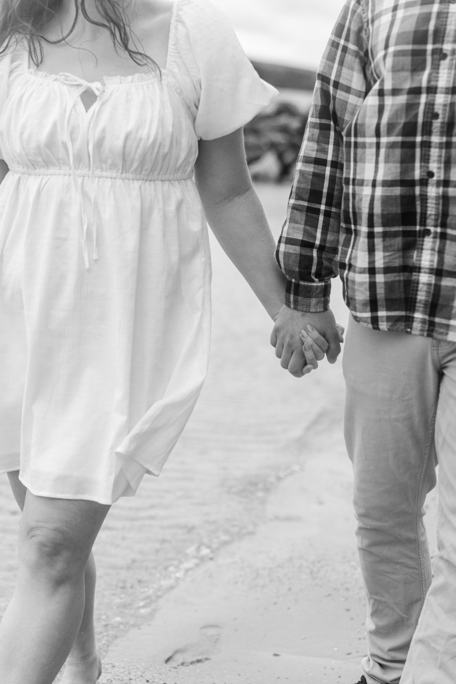 Black and white photo of a couple holding hands while walking on a beach, woman in white dress, man in plaid shirt.