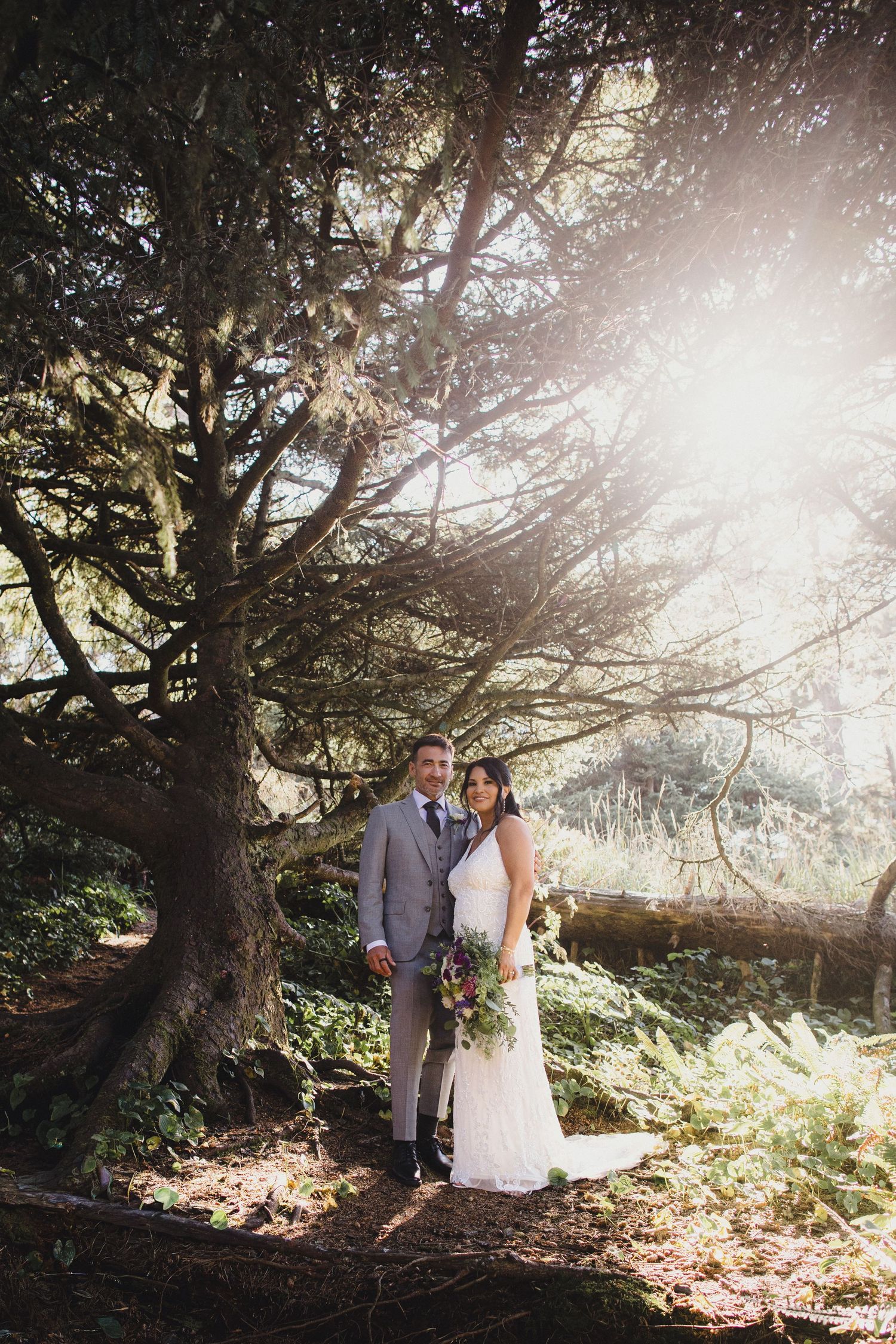 Bride and groom pose under a large tree with sunlight streaming through branches in a lush forest wedding portrait.