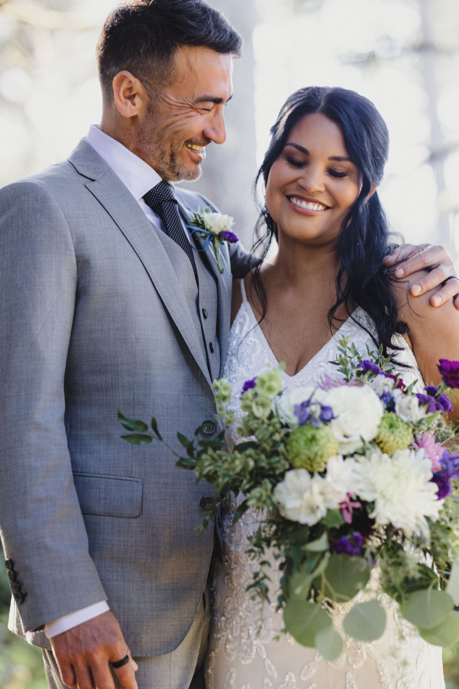 Smiling bride holding purple and white bouquet stands beside groom in gray suit at outdoor wedding ceremony.