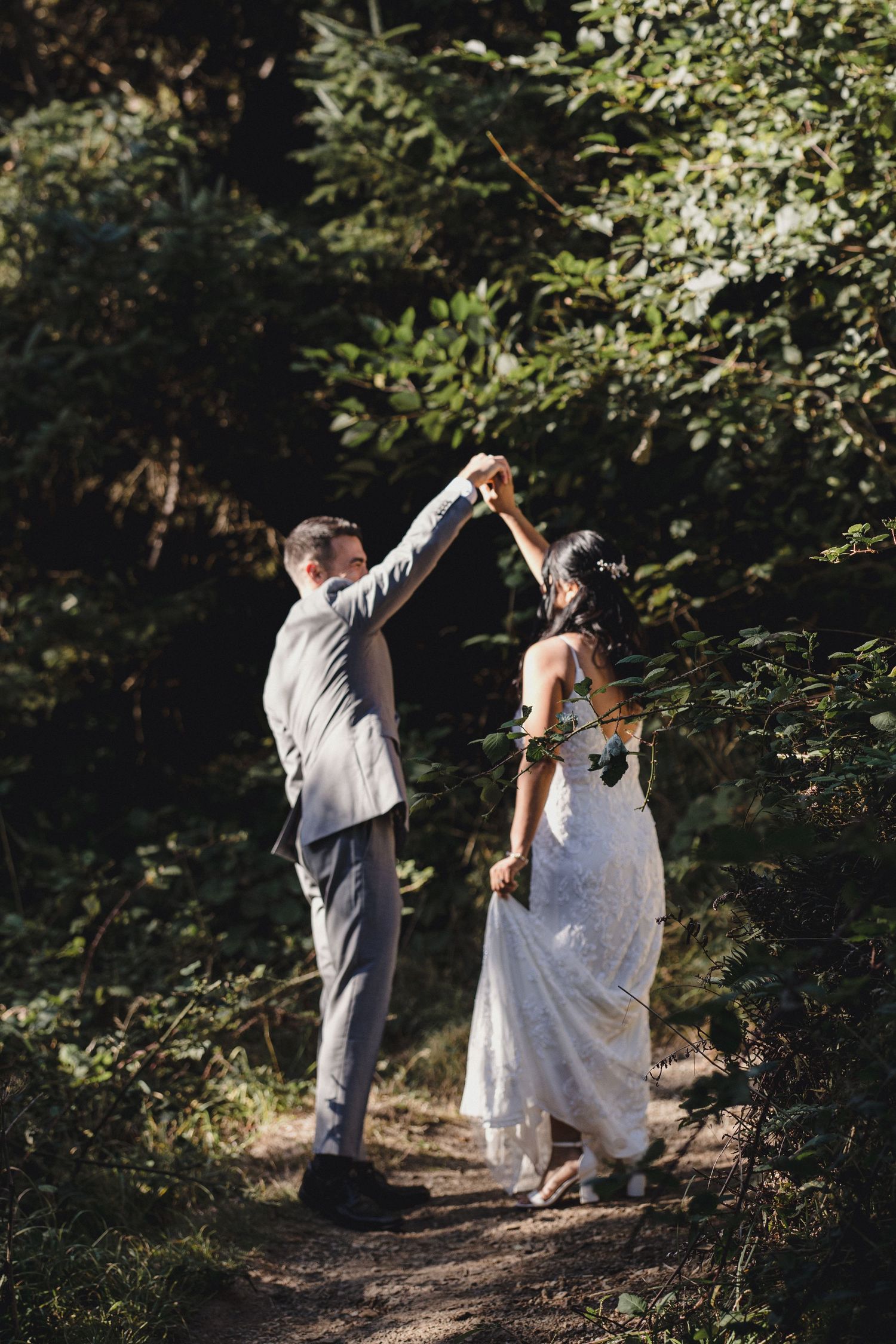 Bride and groom dancing on a forest path, surrounded by lush green trees in soft natural sunlight.