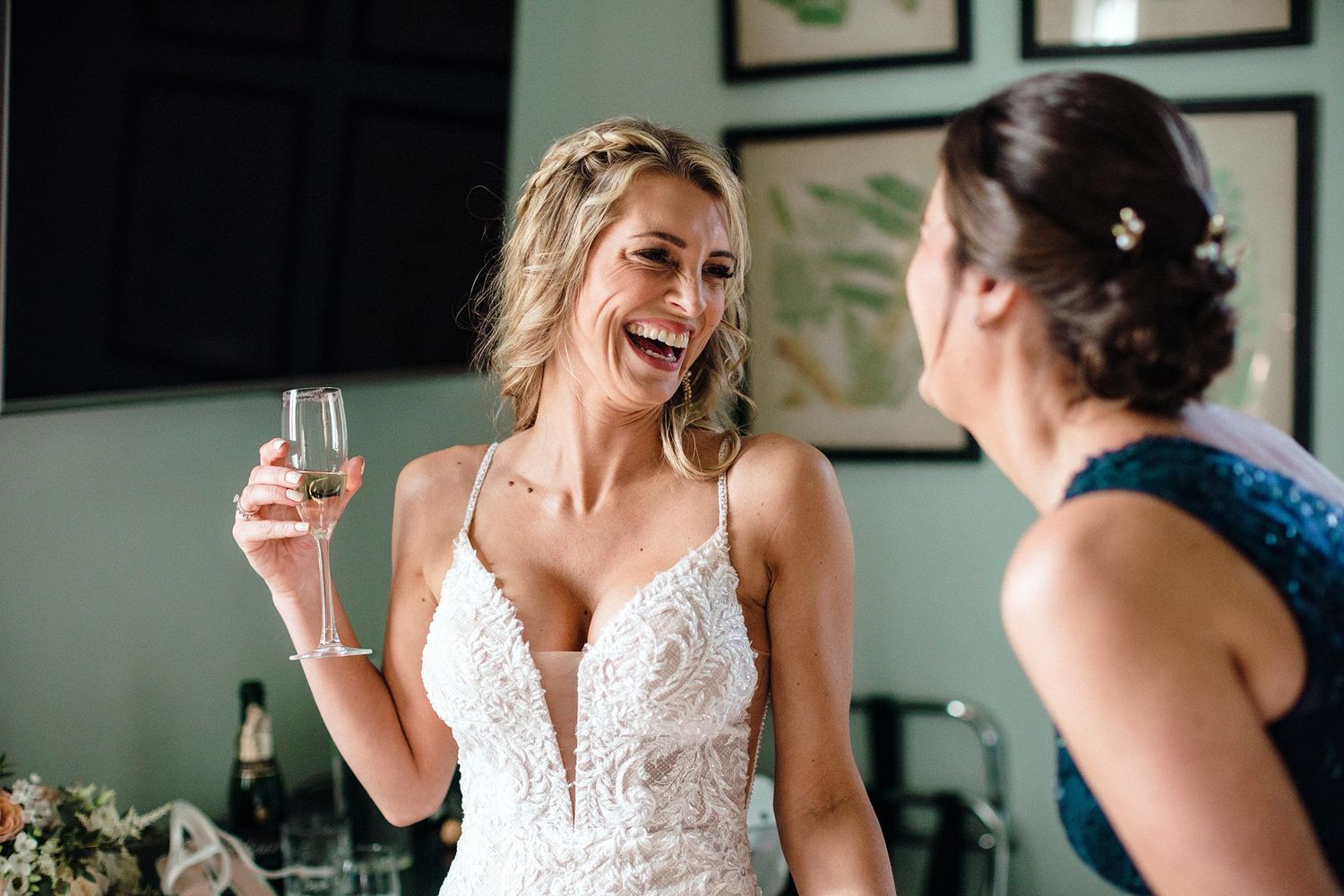 Bride in lace wedding dress laughing and holding champagne glass while talking with bridesmaid before wedding ceremony.