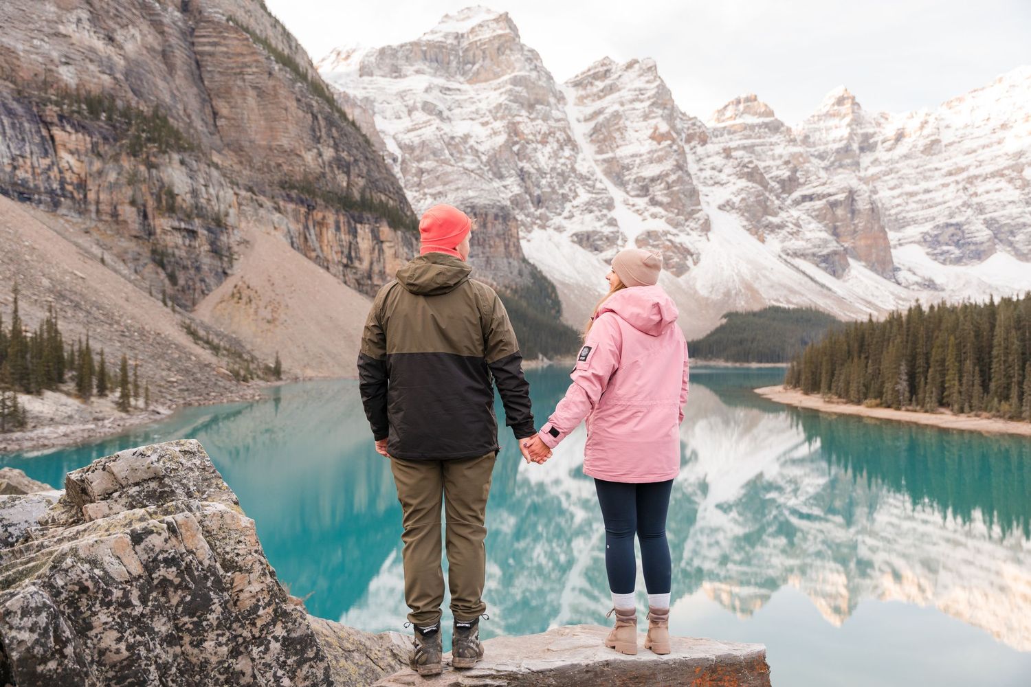 Couple holding hands at Moraine Lake, Banff, with turquoise water and snow-capped Rocky Mountains in background.