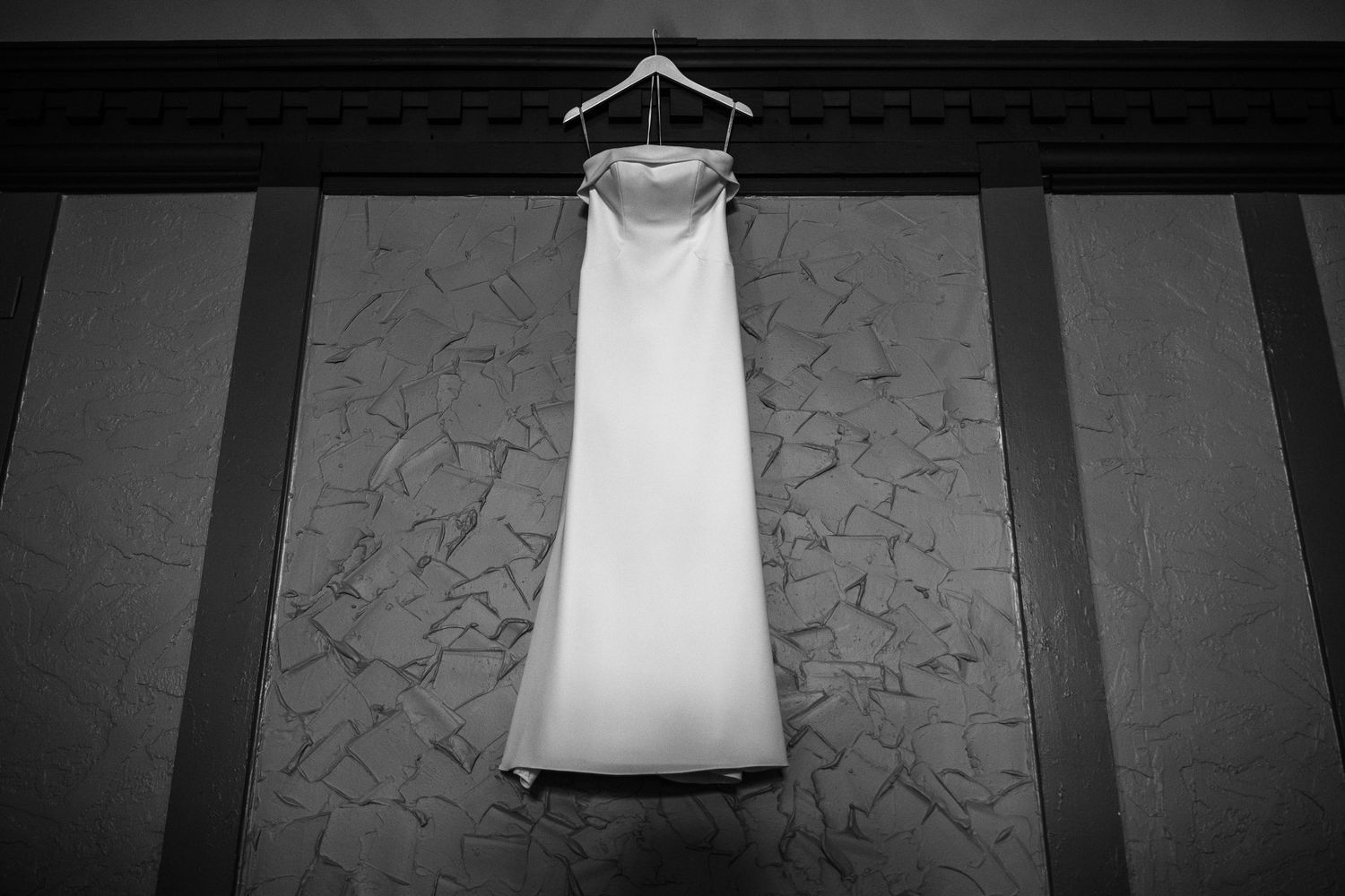 Black and white photo of an elegant strapless wedding gown hanging on a hanger against a textured wall.
