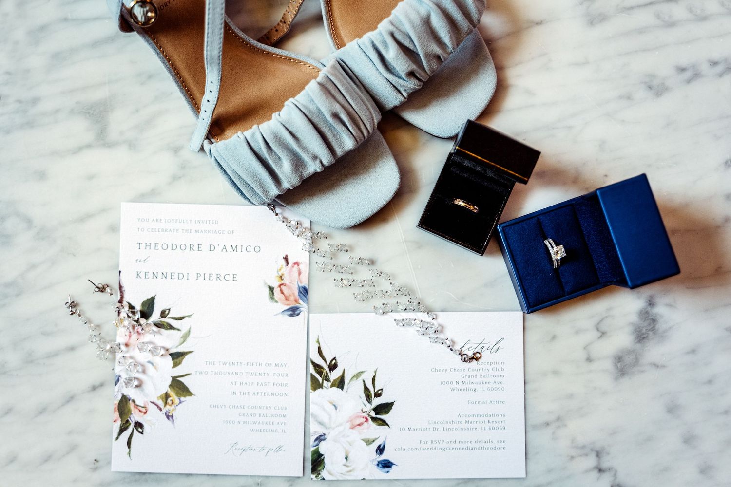 Wedding flat lay with floral invitations, blue heels, diamond rings in boxes on marble surface.