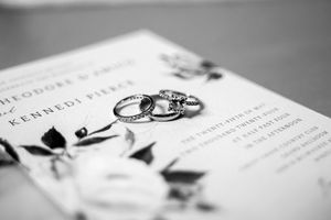 Black and white photo of wedding rings resting on a floral wedding invitation for Theodore and Kennedi Pierce.