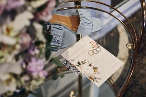 Elegant wedding stationery with floral design displayed on glass table beside bridal shoes and purple flowers.