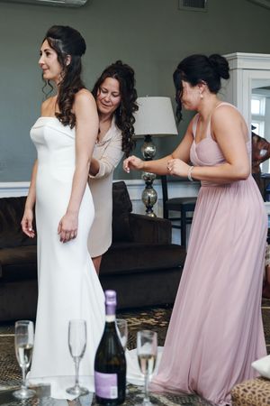 Bride being helped into white wedding gown by bridesmaids in pink dress, champagne glasses on table nearby.