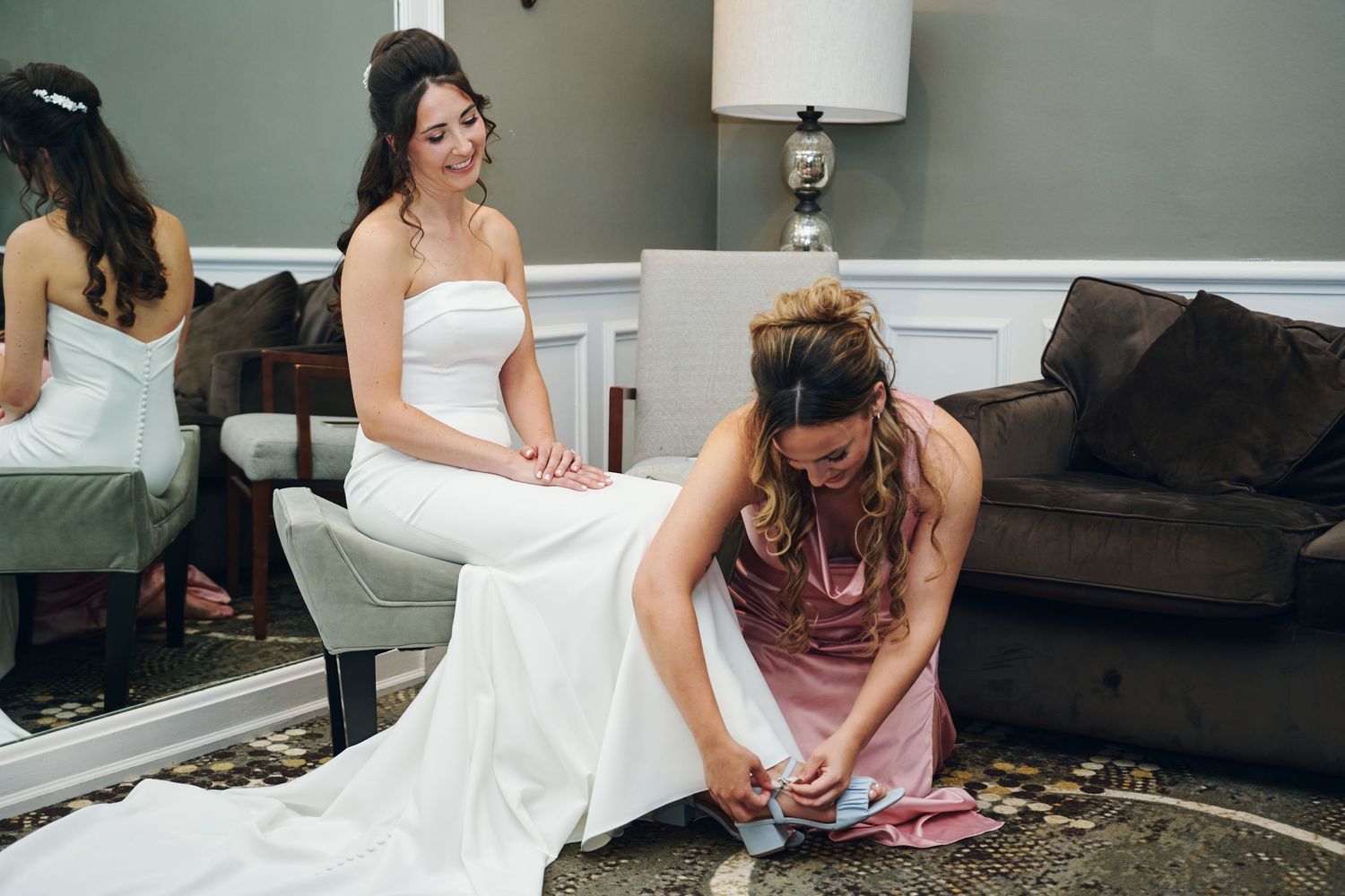 Bridesmaid in pink dress kneeling to help bride with shoes while bride sits in white wedding gown before ceremony.