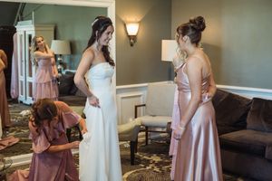 Bride in white gown being helped by bridesmaids in dusty rose dresses in elegant hotel room before wedding ceremony.