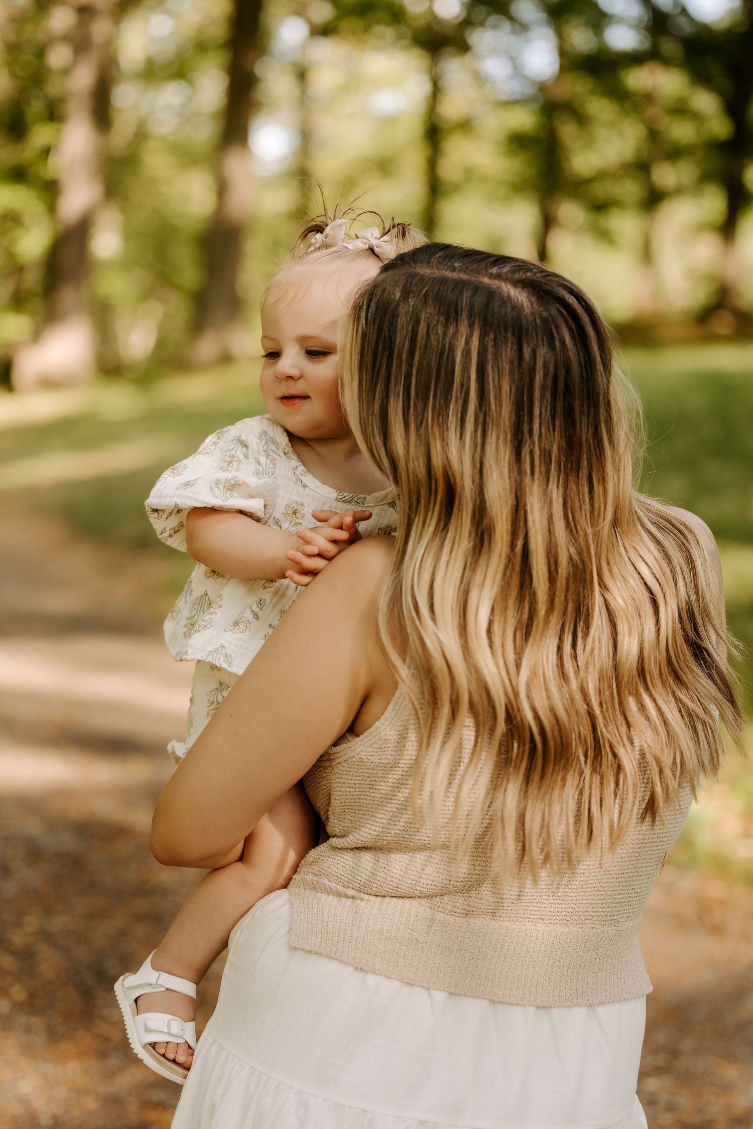 A mother with long blonde hair holds her toddler daughter in a floral dress outdoors in a sunlit park.