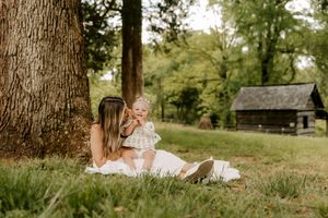 A mother sits against a large tree in a lush green park, lovingly holding her baby in a sunlit outdoor setting.