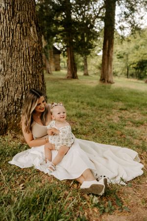 A mother sits against a tree in a white skirt, smiling at her happy baby girl in a floral romper outdoors.