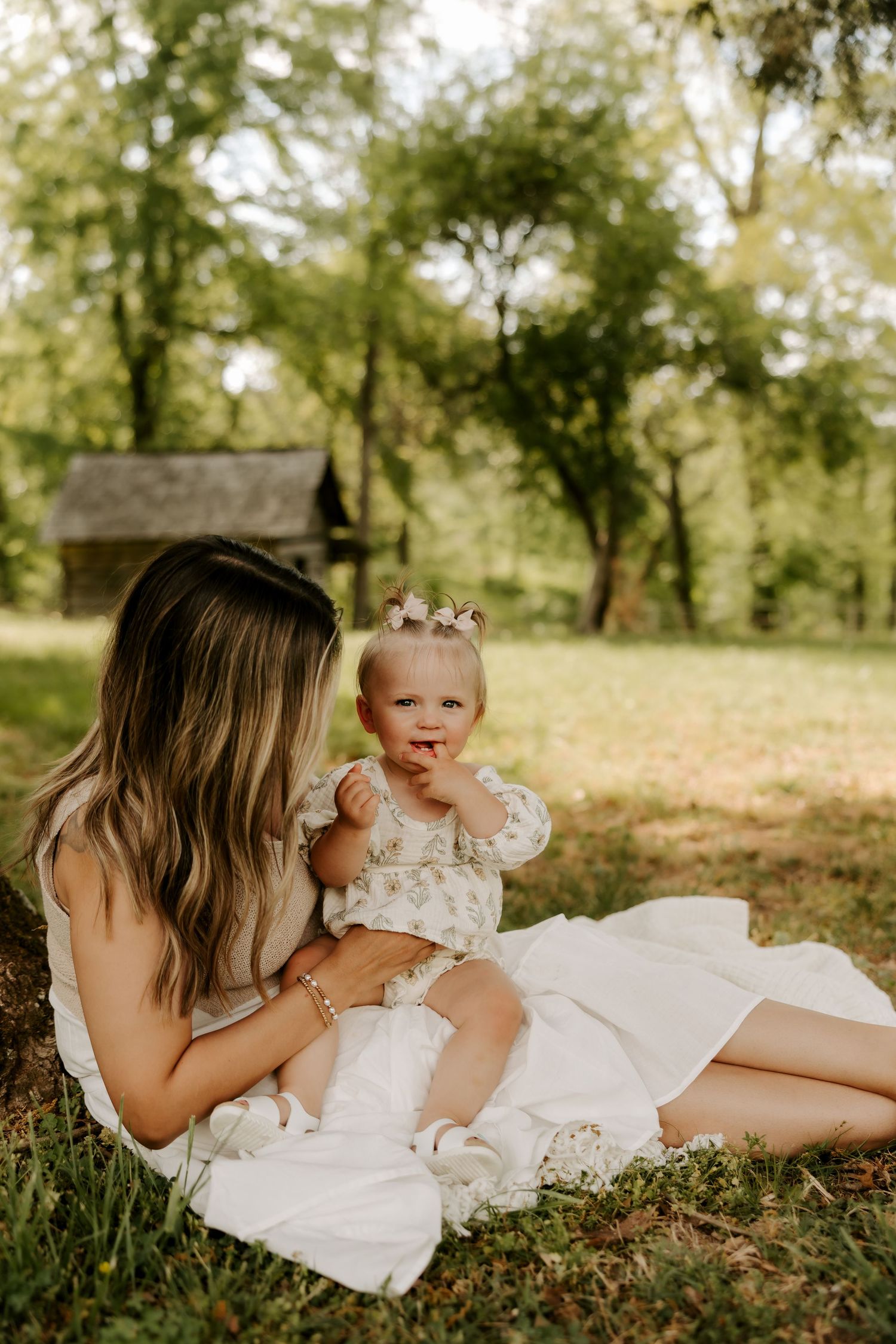 A mother sits on grass holding her baby girl in a sunny park with trees and a rustic wooden structure behind them.