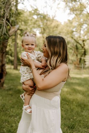 A mother holds her smiling baby girl outdoors in a lush green park, both dressed in light summer outfits.
