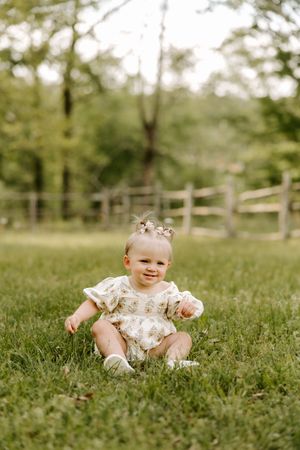 Smiling baby girl in floral romper sitting on green grass outdoors with wooden fence and trees in background.