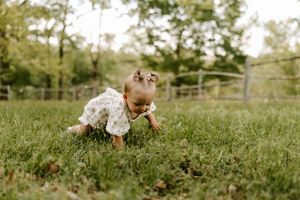 A baby girl in a floral romper crawls on green grass outdoors with trees and a wooden fence in the background.