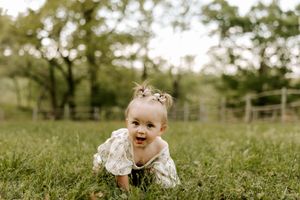 Adorable baby girl in a floral dress crawling on green grass outdoors with trees and a fence in the background.