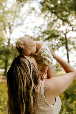 Mother lifting smiling baby girl in floral romper outdoors in sunny green park setting.
