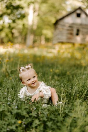 Happy baby girl in floral romper sitting in green grass near rustic barn on sunny day.