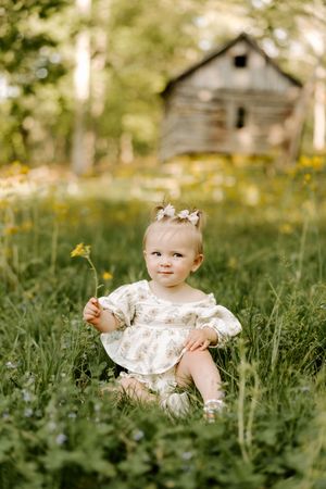 Adorable baby girl in white floral dress sitting in green meadow holding a wildflower near rustic wooden barn.
