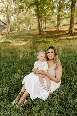 Mother in beige dress sitting in wildflower field holding baby girl in white floral outfit surrounded by green trees.
