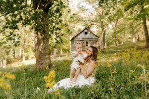 A mother sits in a lush green meadow holding her baby, smiling joyfully, with wildflowers and a rustic barn in the background.