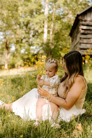 A young mother sits in lush green grass holding her toddler daughter near wildflowers and a rustic wooden barn.