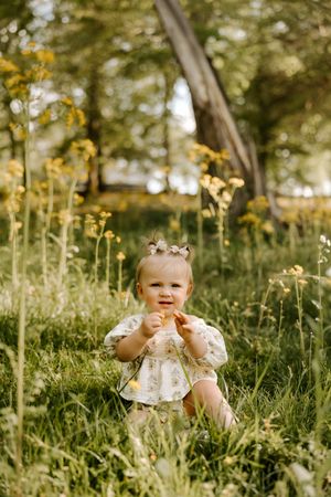 Toddler girl in floral dress sitting in green meadow with yellow wildflowers on sunny day.