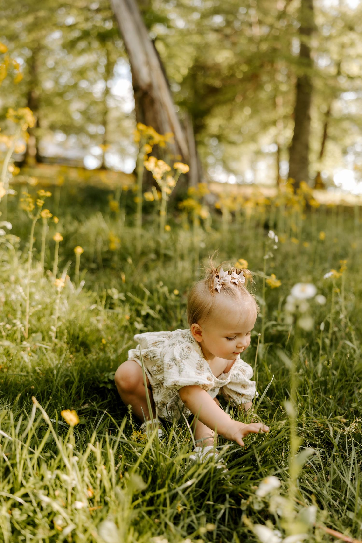 A baby girl in a floral romper explores a sunny meadow with wildflowers and green grass.