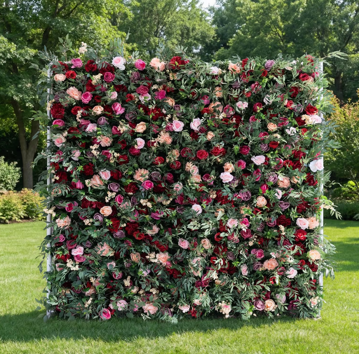 Lush outdoor floral wall backdrop with red, pink and white roses surrounded by green foliage.